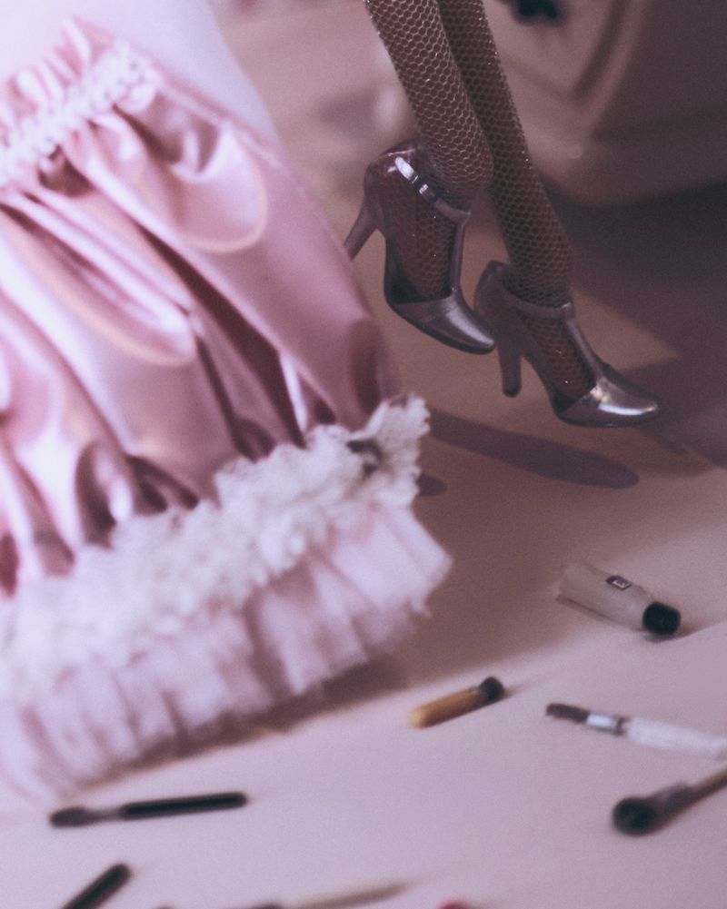 Close-up of doll's legs in high heels with a pink frilly skirt and makeup brushes scattered on a pink surface.