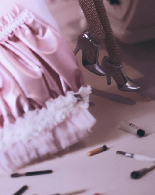 Close-up of doll's legs in high heels with a pink frilly skirt and makeup brushes scattered on a pink surface.
