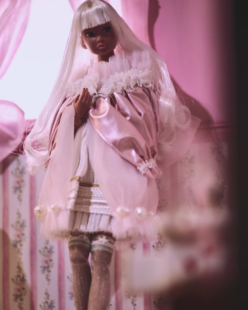 Fashion doll with fair skin and long white hair wearing a pink and white dress with lace details posing indoors against a pink backdrop.