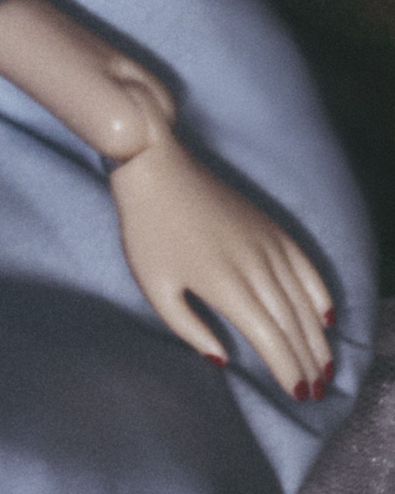 Close-up of an adult woman's arm and hand with red nail polish, in a soft, blurred indoor setting with blue and gray fabrics.