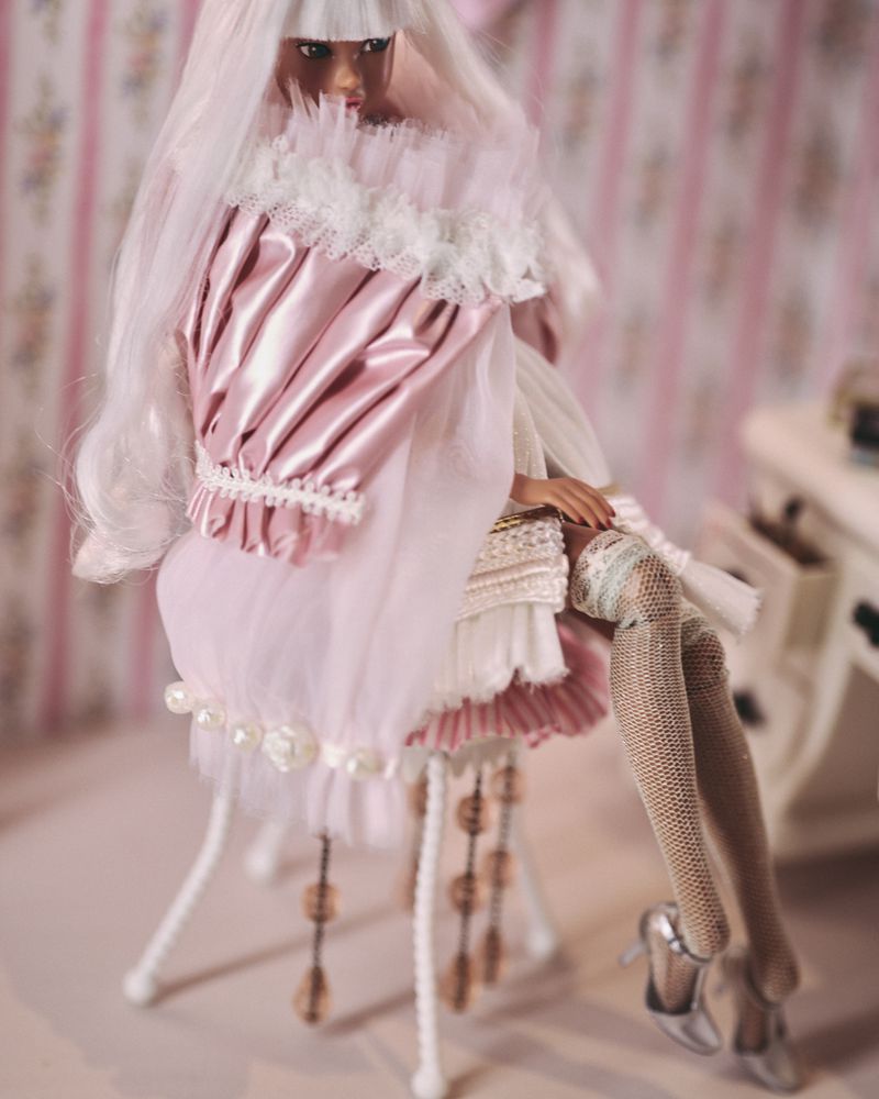 Seated fashion doll with long white hair, wearing a pink frilly dress and silver heels, in front of a patterned wall and table.