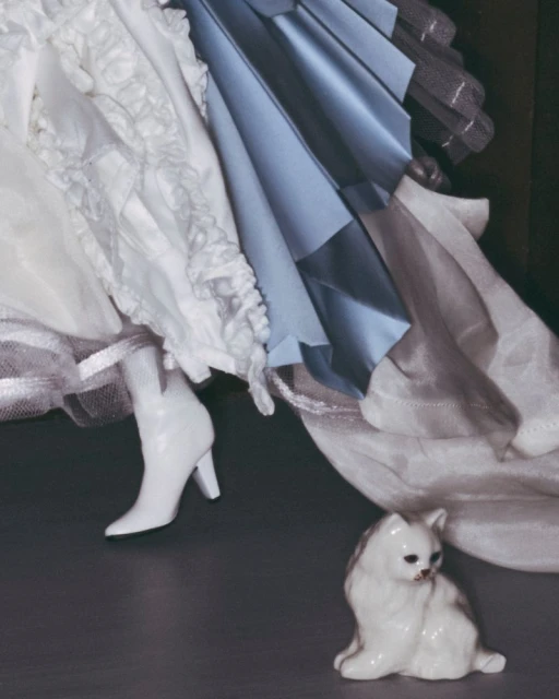 Fashion shot of a woman's legs in white heeled boots, a white dress, and a ceramic cat figurine on a dark background. 