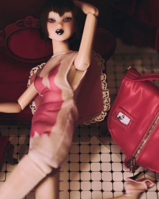 A fashion doll with bangs and a pink dress poses next to a pink purse on a checkered surface with a red chair visible in the background.