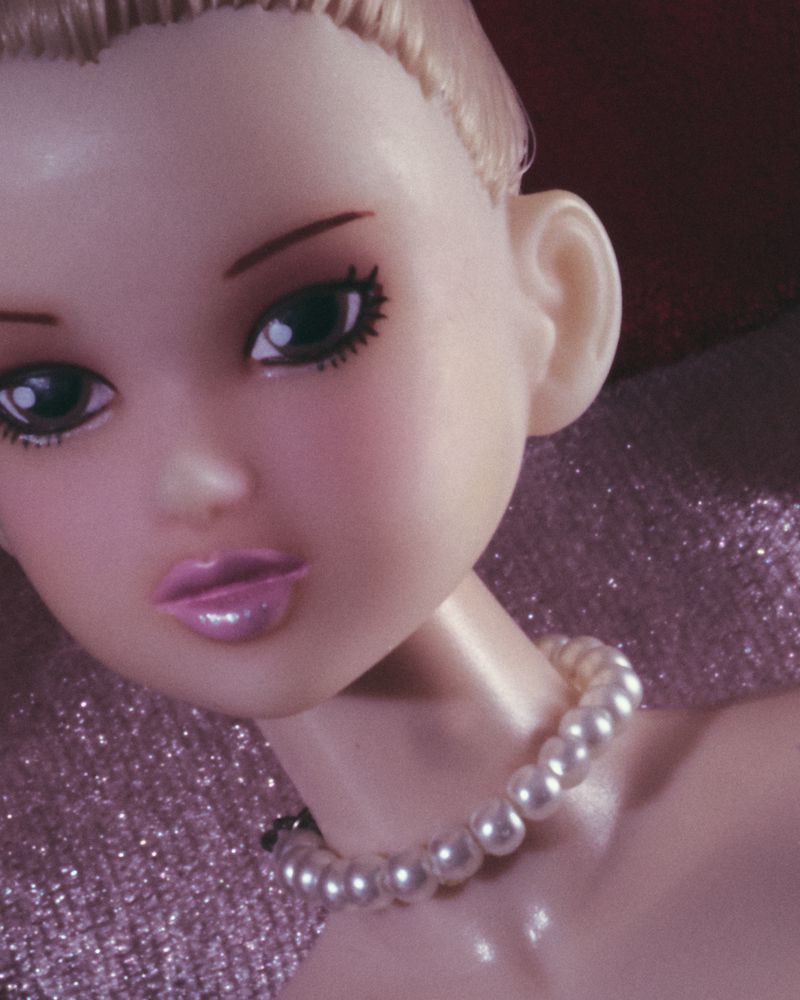 Close-up of a fashion doll's face with pearl necklace and blond hair against a shimmering pink backdrop in a beauty or fashion context.