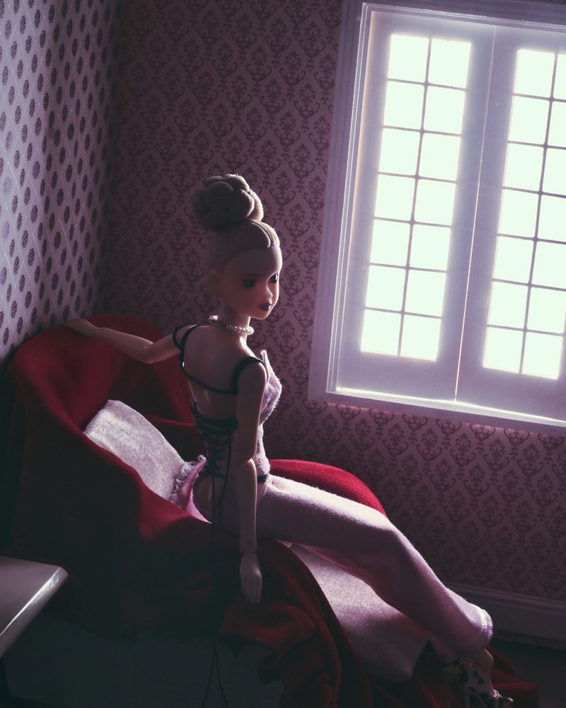Articulated doll sitting on a red chaise lounge in front of a window with a patterned wallpaper background and neutral light.