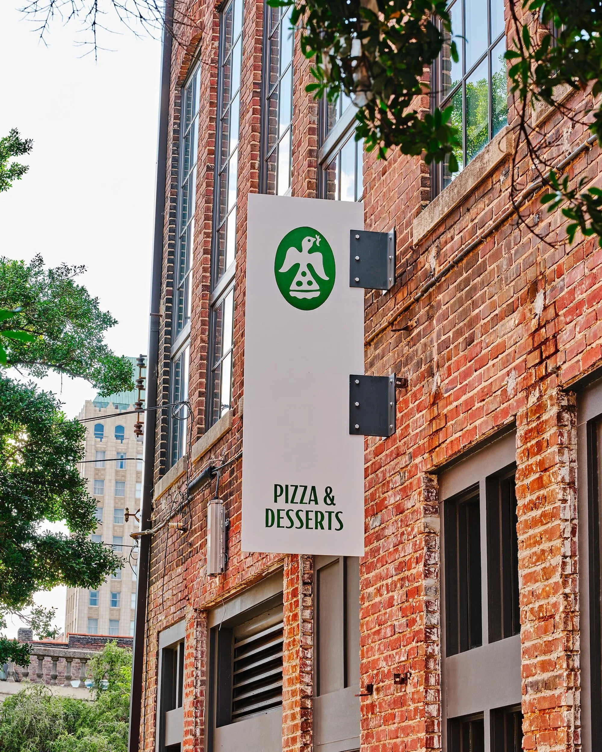 Outdoor view of a white restaurant sign on a brick building featuring a green logo, and the words, Pizza & Desserts.