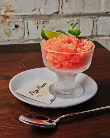 Frozen dessert with lime and mint garnish in glass bowl on a plate with spoon, atop a wooden table with a brick wall backdrop.