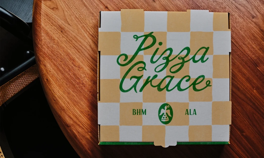 Overhead shot of a pizza box with a checkered pattern and the words Pizza Grace on a wooden table, with a chair visible.