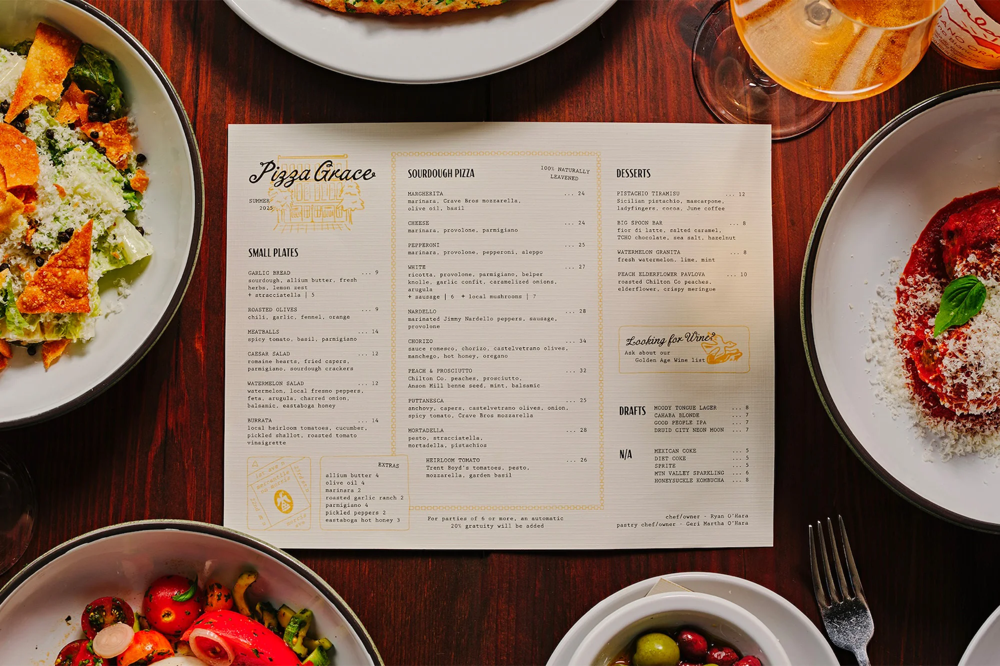 Overhead shot of a Pizza Grace menu surrounded by plates of salad, tomatoes and burrata, and pasta on a dark wooden table.
