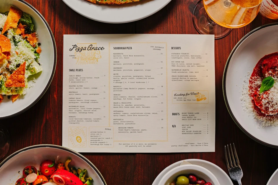 Overhead shot of a Pizza Grace menu surrounded by plates of salad, tomatoes and burrata, and pasta on a dark wooden table.