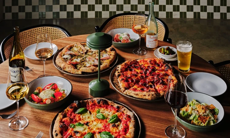 Overhead shot of a wooden table filled with pizzas, salads, wine, and beer in a restaurant setting with patterned green and white tiles.