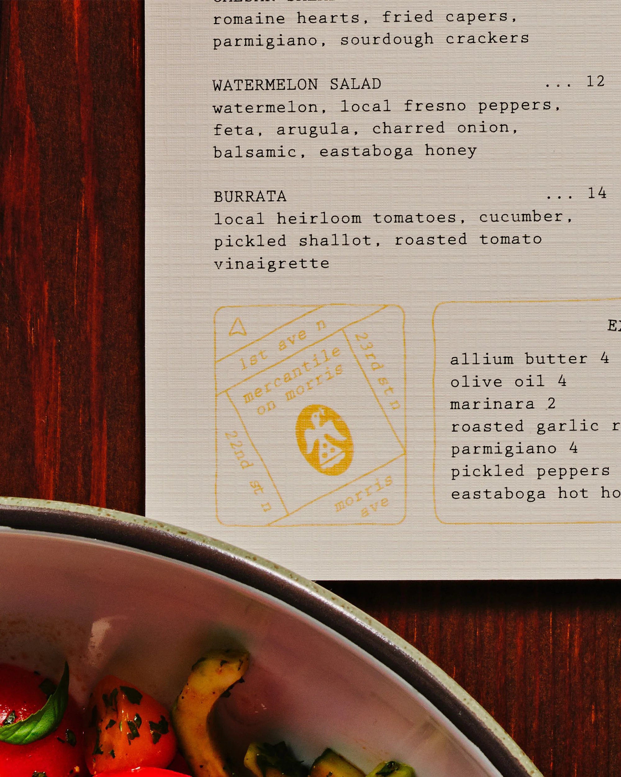 Overhead view of a restaurant menu featuring dishes like watermelon salad and burrata, complemented by a bowl of salad.