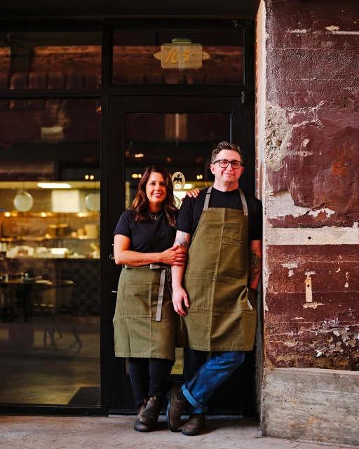 Two restaurant owners, a woman and a man, stand in front of their restaurant, wearing aprons and smiling, showcasing their establishment.