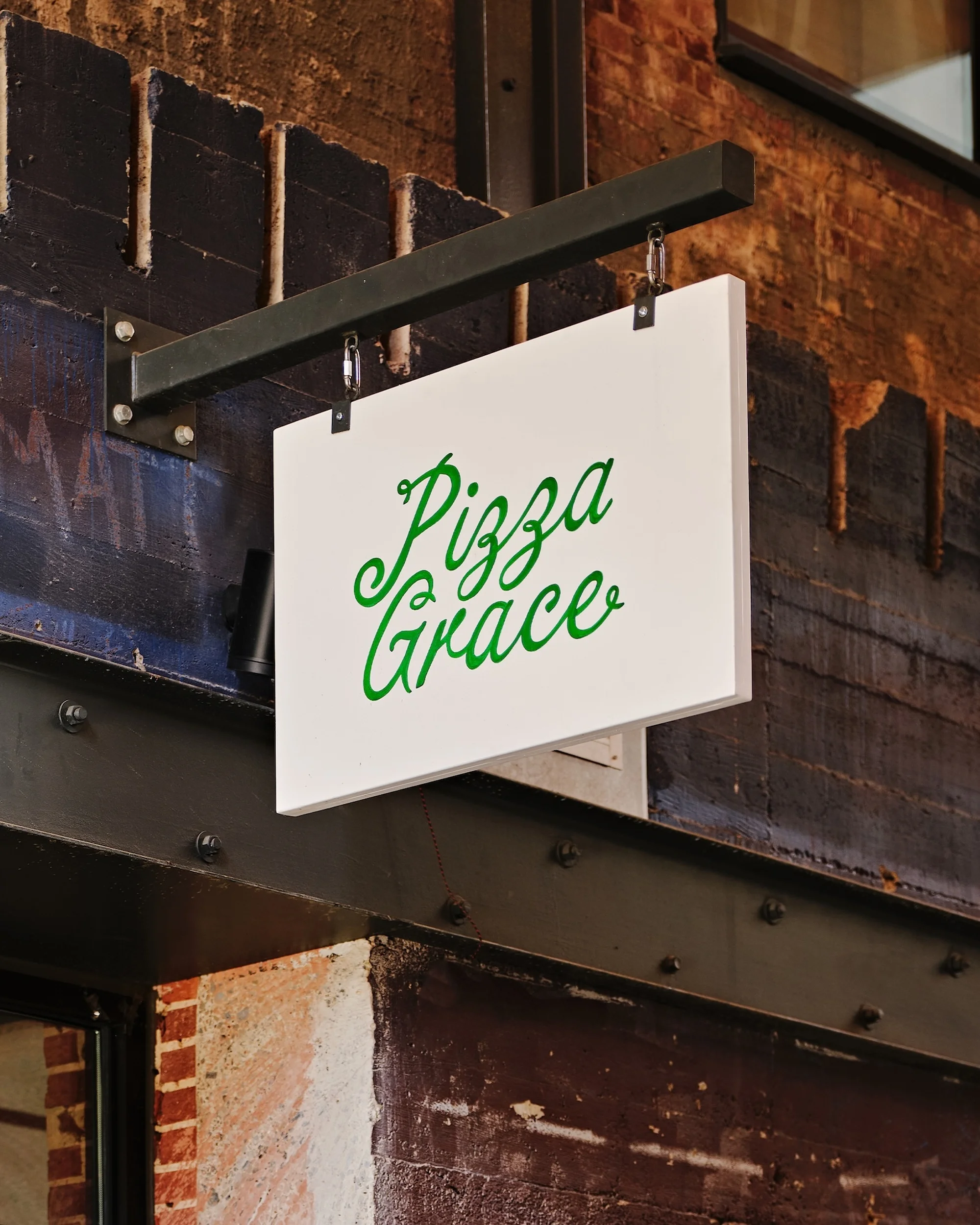 White rectangular hanging storefront sign displaying green cursive lettering for Pizza Grace against a dark brick and metal wall