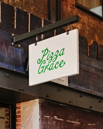White rectangular hanging storefront sign displaying green cursive lettering for Pizza Grace against a dark brick and metal wall