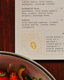 Close-up of a textured menu sheet with typographic text next to a ceramic bowl of salad on a wooden table.