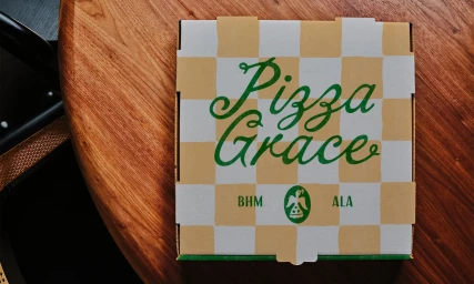 Square pizza box with a yellow and white checkered pattern and green script typography reading Pizza Grace resting on a wooden surface.