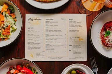 Overhead view of a restaurant menu surrounded by plates of salads and pizza on a dark wooden table.