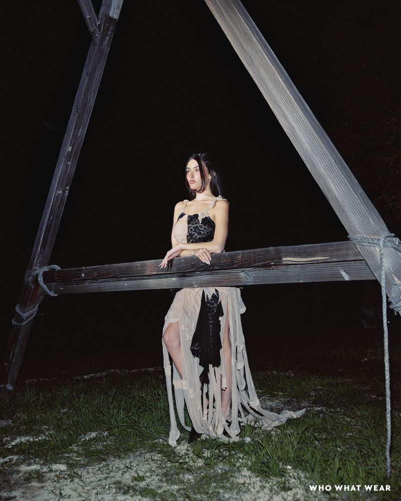 Woman in a flowing beige gown with black lace accents poses outdoors at night, leaning against a wooden structure.