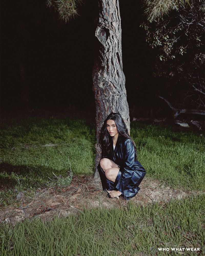 Madison Beer poses in a black leather jacket and shorts, kneeling next to a large tree on grassy ground in an outdoor setting.