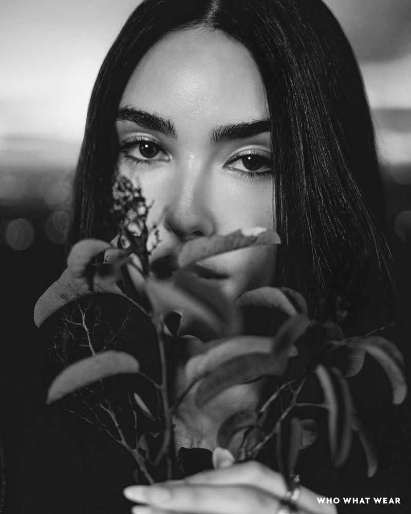 Black and white portrait of a woman with dark hair partially obscuring her face with leafy plants against a blurred background.