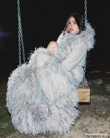 Madison Beer seated on a wooden swing at night wearing a voluminous feathery grey coat for a Who What Wear feature