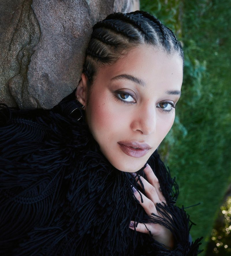 Portrait of a young woman with braided hair wearing a black fringed top, posing against a gray rock and green foliage backdrop.