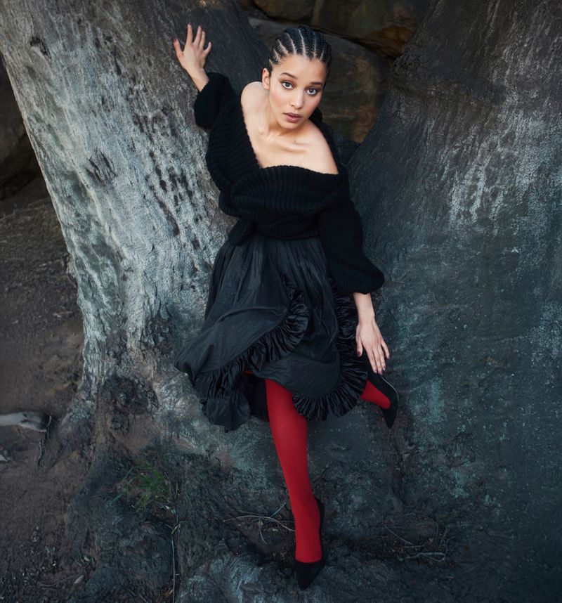 Fashionable woman poses against a rock wearing a black sweater, ruffled skirt, red tights, and high heels in an outdoor environment.
