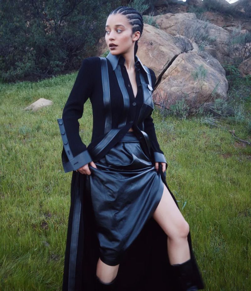 Woman in a black outfit with a leather skirt, standing in a grassy field with rocks in the background, looking off to the side.