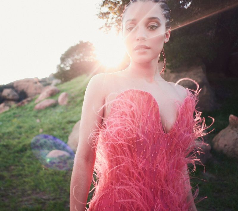 A woman in a pink feather dress poses in a sunlit outdoor setting with greenery and rocks in the background, creating a glamorous and natural portrait.