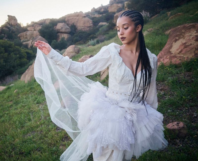 A woman poses outdoors in a white dress with a tulle skirt, set against a rocky landscape with greenery at daytime.