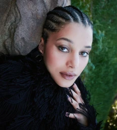 Portrait of Chase Infiniti wearing a black fringed garment, leaning against a textured stone wall outdoors.