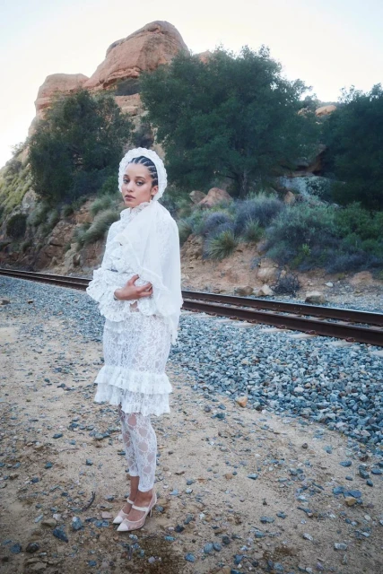 Chase Infiniti wearing a white lace dress and headpiece standing by railroad tracks for The Sunday Times Style Magazine UK
