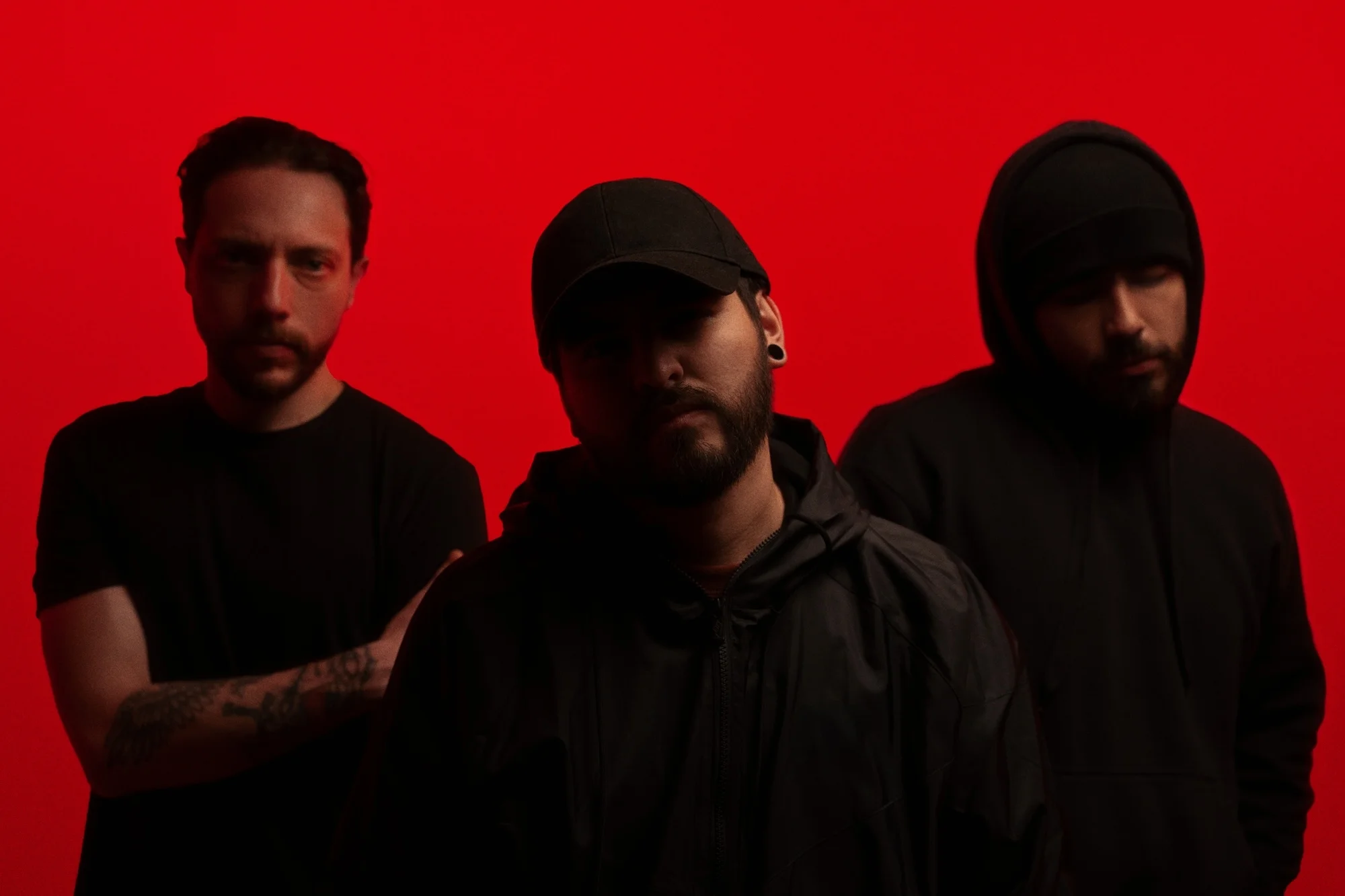 Three adult men stand in a group against a red backdrop, all dressed in black tops, with one wearing a baseball cap.