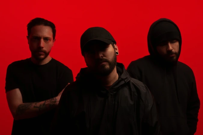Three adult men stand in a group against a red backdrop, all dressed in black tops, with one wearing a baseball cap.