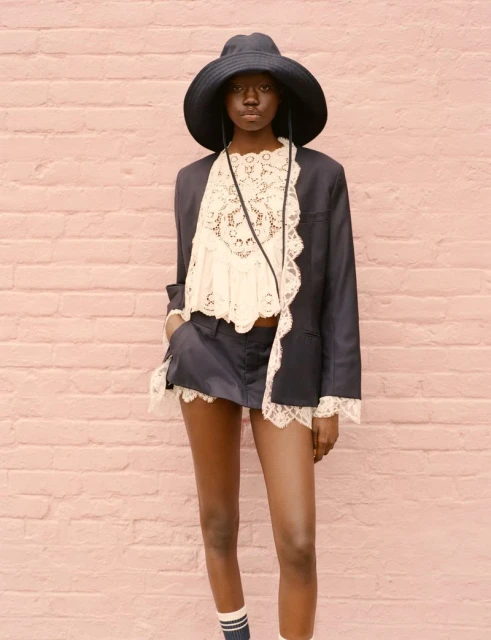 A Black woman poses in a navy shorts suit with lace trim and a wide-brimmed hat against a pink brick wall, styled for high fashion.