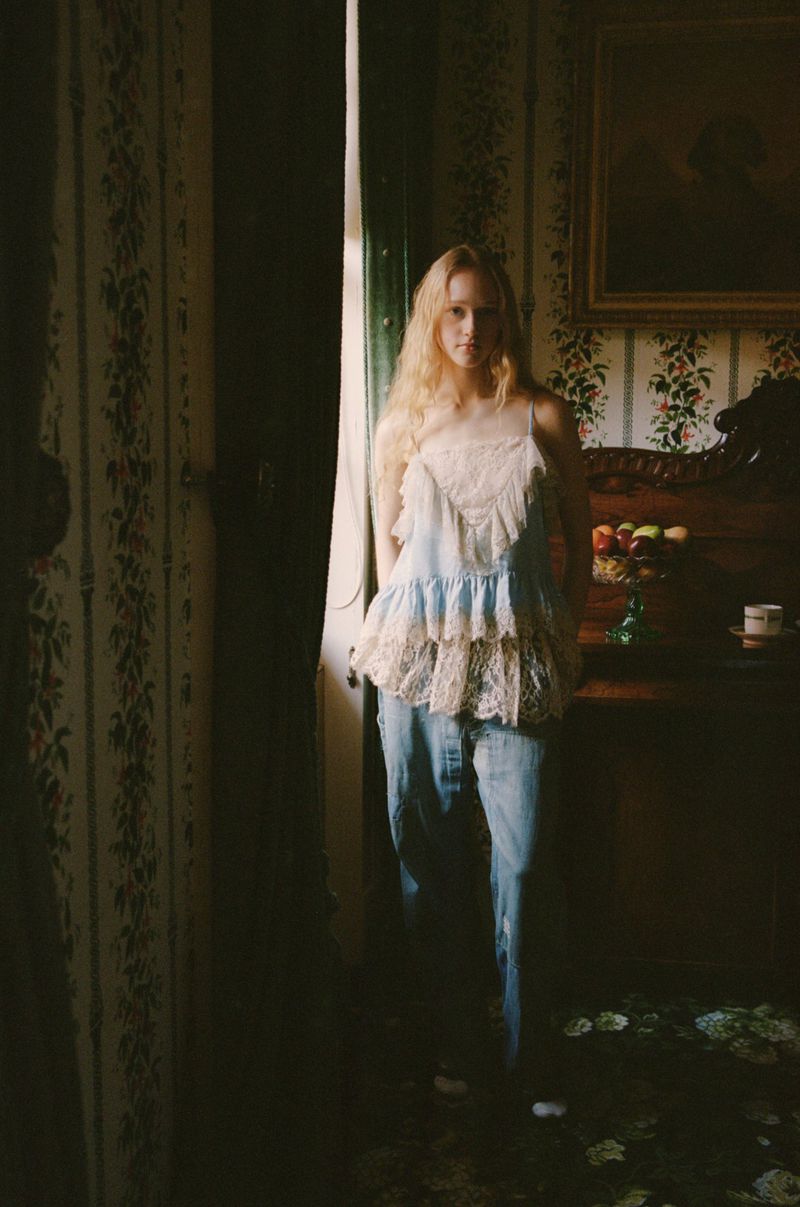 Woman with long blonde hair poses in blue and white lace top and jeans in a room with patterned wallpaper and dark wood furniture.