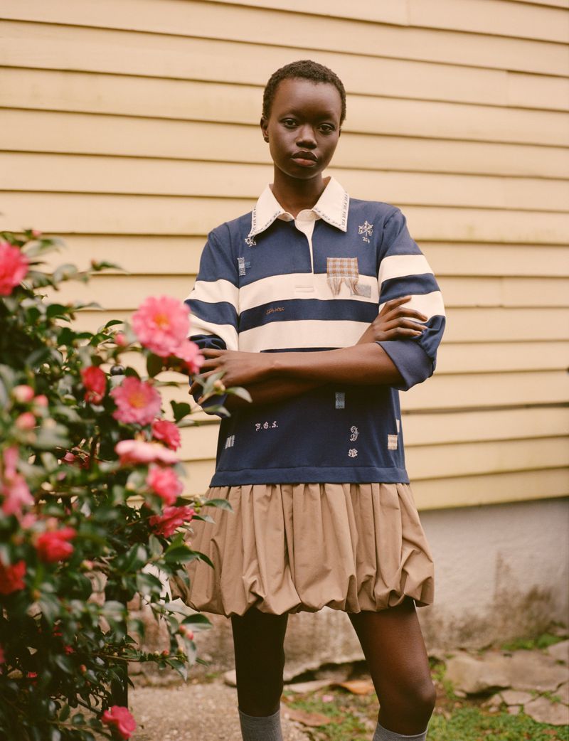 A fashionable young woman with short hair stands in front of a tan siding wall with pink flowers near the camera, looking at viewer.