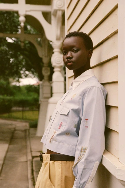A young Black woman with short hair leans against a white house wearing a light blue shirt and tan pants in an outdoor setting.