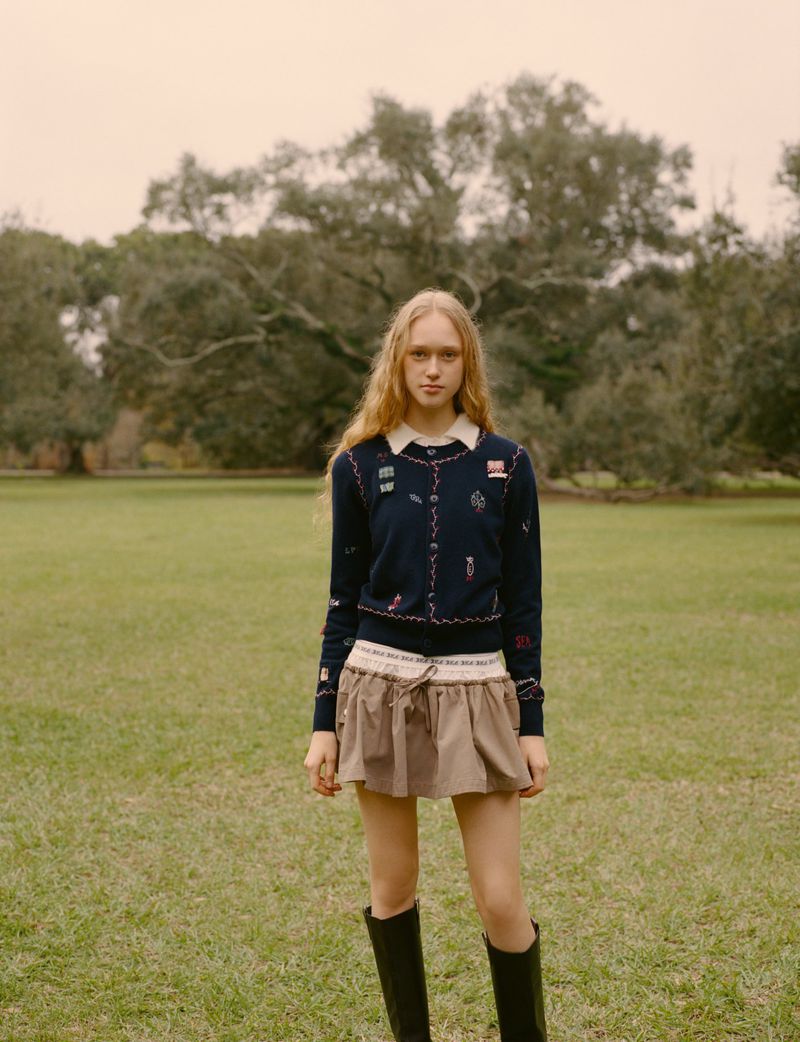 Blonde woman stands in a grassy field, wearing a navy cardigan with patches, a neutral miniskirt, and black boots, with trees behind her.