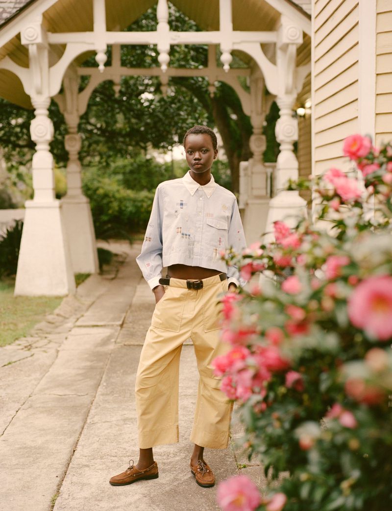 A slender woman with short dark hair is standing in front of a lush garden with a white pergola in the background on a sunny day.