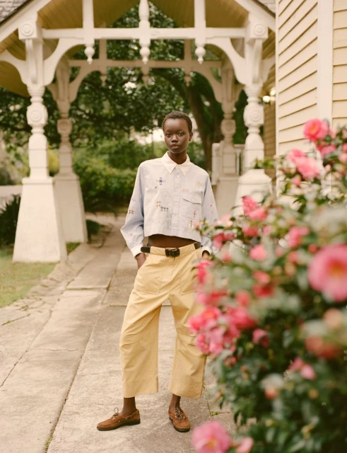 A slender woman with short dark hair is standing in front of a lush garden with a white pergola in the background on a sunny day.
