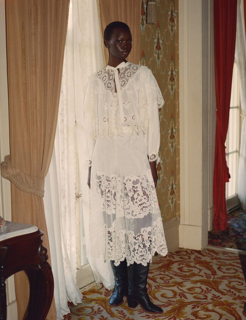 A Black female fashion model showcases a long white lace dress, matching capelet, and black boots in a room with patterned wallpaper.