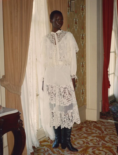A Black female fashion model showcases a long white lace dress, matching capelet, and black boots in a room with patterned wallpaper.
