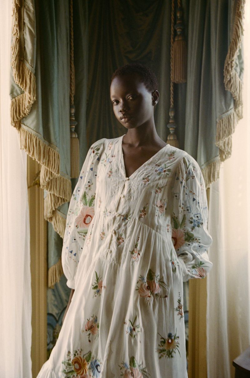 A woman wearing a floral-patterned dress poses indoors in front of draped curtains, showcasing fashion and elegant portraiture.