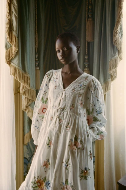 A woman wearing a floral-patterned dress poses indoors in front of draped curtains, showcasing fashion and elegant portraiture.