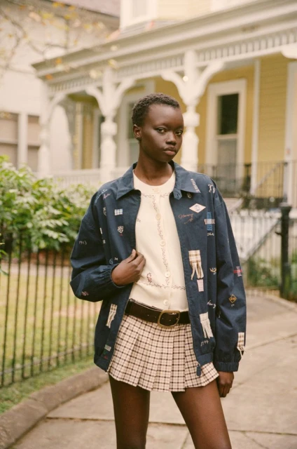 Young African woman poses outdoors wearing a patterned jacket, cream sweater, and plaid pleated skirt against a yellow house backdrop.