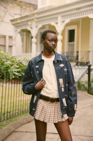 Young African woman poses outdoors wearing a patterned jacket, cream sweater, and plaid pleated skirt against a yellow house backdrop.