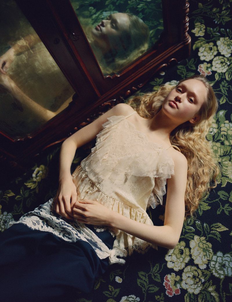 Young woman with long blonde hair lies on a floral couch next to a vintage mirror in an indoor setting, wearing a white lace top.