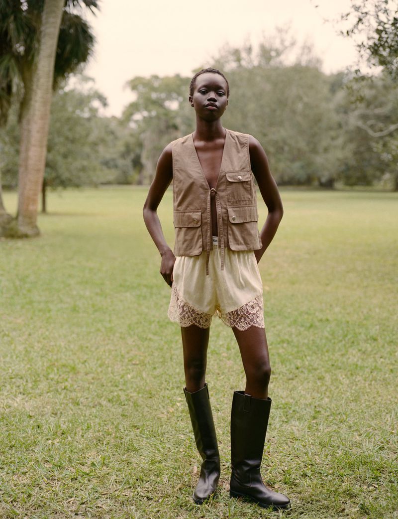 A slender woman with dark skin stands outdoors in riding boots, lace shorts, and a vest against a verdant background.